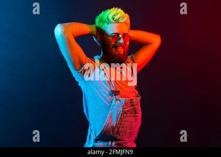 Attraktiver junger, erwachsener Mann mit positivem Gesichtsausdruck, der mit einem charmanten Lächeln auf die Kamera blickt, die Hände hinter dem Kopf hochhebt und Overalls trägt. Stockfoto