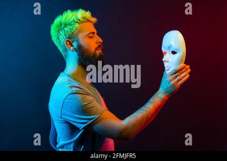 Seitenansicht eines selbstbewussten ernsthaften Mannes in Denim-Overalls, der mit aufmerksamem Blick die weiße Maske in den Händen hält und anschaut und versucht, Verstecke zu verstehen Stockfoto