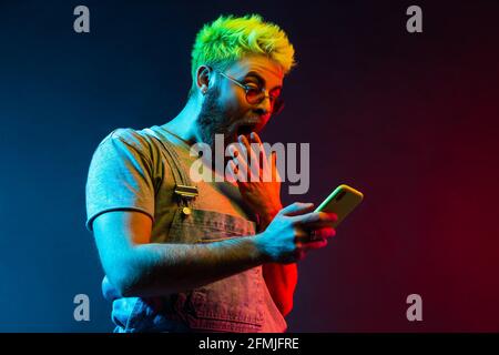 Junger Erwachsener attraktiver Hipster Kerl mit Bart mit Handy in der Hand sieht etwas Erstaunliches auf dem Display, bedeckt weit geöffneten Mund mit Handfläche. C Stockfoto