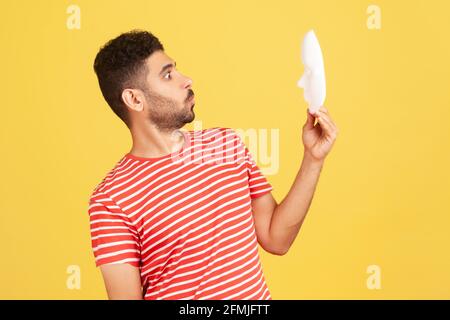 Profilportrait eines bärtigen Mannes im gestreiften T-Shirt, der auf eine weiße Maske mit einem unbekannten Gesicht mit überraschtem Ausdruck schaut und versucht, Emotionen zu erraten. Innen Stockfoto