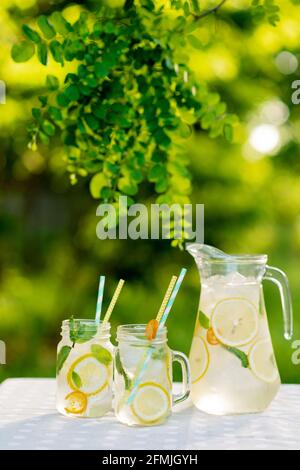 Limonade-Erfrischungsgetränk in einem Krug und Gläser mit Zitronen, frischer Minze und Eis auf einem Gartentisch. Picknick im Sommer im Freien. Weicher, selektiver Fokus. Stockfoto