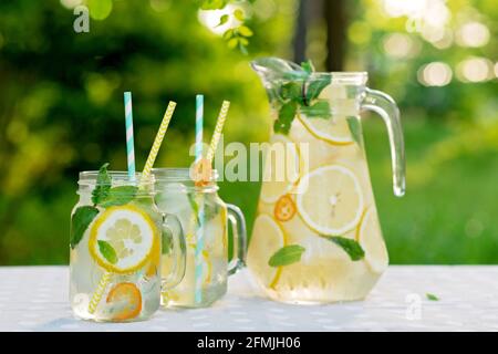 Limonade-Erfrischungsgetränk in einem Krug und Gläser mit Zitronen, frischer Minze und Eis auf einem Gartentisch. Picknick im Sommer im Freien. Weicher, selektiver Fokus. Stockfoto