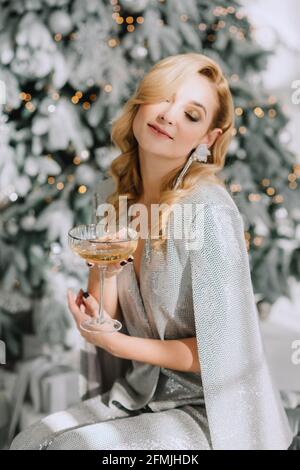 Junge schöne luxuriöse blonde Frau mit einem Glas in ihr Hands feiert das neue Jahr vor dem Hintergrund von Unschärfe Lichter Bokeh von Weihnachten Stockfoto