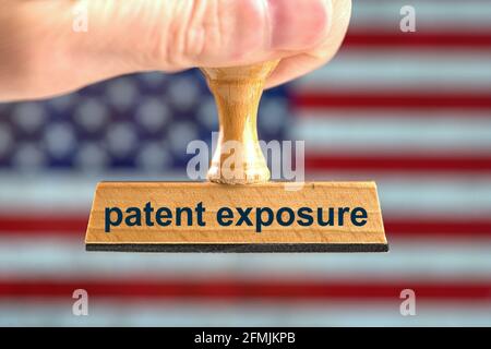 Deutschland. Mai 2021. Ein symbolischer Holzstempel mit der Aufschrift „Patent Exposure“, der von einer Hand im Bluten vor einer verschwommenen US-Flagge im Hintergrund gehalten wird. Die USA setzen die Patente für Corona-Impfstoffe aus. Foto mit Komposition [M]. Credit: dpa/Alamy Live News Stockfoto