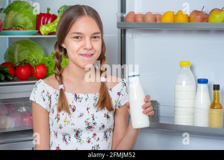 Schöne junge Teenager-Mädchen hält Flasche Milch und Getränke Während Sie in der Nähe des offenen Kühlschranks in der Küche zu Hause stehen Stockfoto