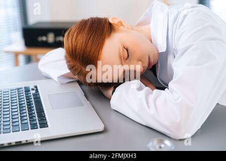 Nahaufnahme einer müden, überarbeiteten Rotschopf-Ärztin im weißen Mantel Schlafen am Schreibtisch mit Laptop und Stethoskop am Arbeitsplatz in der Nähe Angezeigt Stockfoto