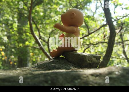 Gruselige Babypuppe, die auf einem Felsen in den Wald sitzt Stockfoto