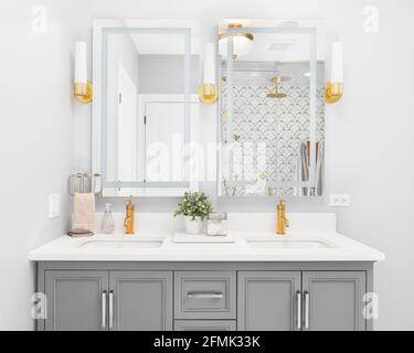 Ein helles, modernes Badezimmer mit grauem Schminkschrank, goldenen Armaturen und Lampen, Granitablagen und Blick auf eine wunderschöne geflieste Dusche. Stockfoto