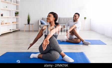 Hübsche junge schwarze Frau macht Twist Yoga Asana mit ihrem Freund zu Hause, Panorama. Millennial African American paar gemeinsam trainieren drinnen d Stockfoto