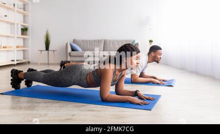 Aktives schwarzes Paar, das zu Hause in der Ellenbogenplanke auf Yogamatten steht, Panorama mit freiem Platz. Afroamerikanische Dame und ihr Freund tun Stärke wor Stockfoto