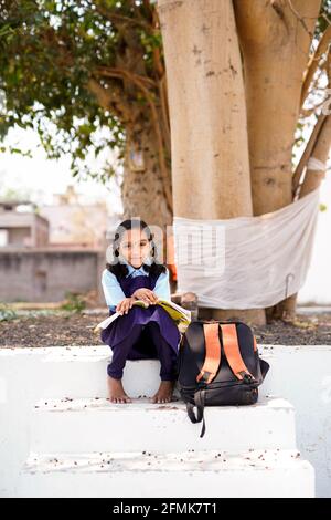 Lächelnde Schülerin trägt Schulrucksack und hält Übungsbuch. Porträt des glücklichen asiatischen jungen Mädchens außerhalb der Grundschule. Nahbereich von Stockfoto