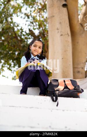 Lächelnde Schülerin trägt Schulrucksack und hält Übungsbuch. Porträt des glücklichen asiatischen jungen Mädchens außerhalb der Grundschule. Nahbereich von Stockfoto