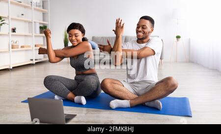 Junges schwarzes Paar, das Yoga oder Pilates praktiziert, in Lotuspose sitzt und während des Haustrainings ihre Arme vor dem Laptop streckt, Panorama. Spo Stockfoto