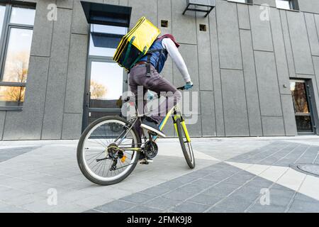 Junger Kurier in medizinischer Maske liefert Lebensmittel mit gelbem Thermalrucksack, fährt mit dem Fahrrad in der Stadt. Konzept für den Lieferservice von Lebensmitteln Stockfoto