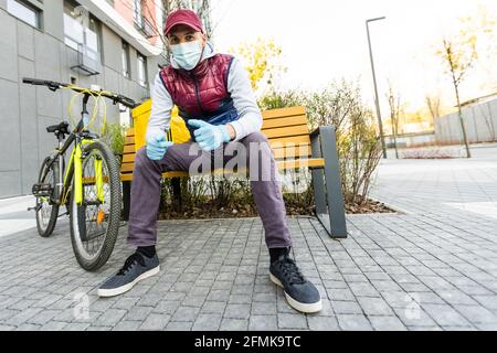 Junger Kurier in medizinischer Maske liefert Lebensmittel mit gelbem Thermalrucksack, fährt mit dem Fahrrad in der Stadt. Konzept für den Lieferservice von Lebensmitteln Stockfoto