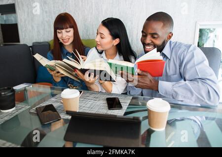 Drei junge aufgeregte multiethnische Freunde, zwei einsame und ein Mann, Studenten, Leser, Literaturliebhaber, die Bücher lesen, die an einem Glastisch im modernen Café sitzen. Konzept für Buchliebhaber Stockfoto