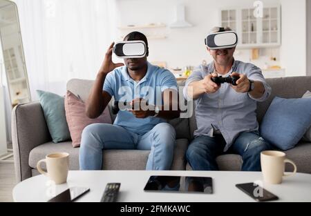 afroamerikanischer Mann und kaukasischer Mann ruhen sich im Zimmer auf dem Sofa aus und spielen Videospiele mit Joysticks. Freunde, die zu Hause Spaß haben. Gam Stockfoto