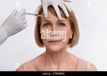 Charmante reife Frau bekommen Schönheit Gesichts-Injektionen von Kosmetologe, Licht Studio Hintergrund. Ältere Dame, die ästhetische Medizin verwendet, um AG zu verhindern Stockfoto