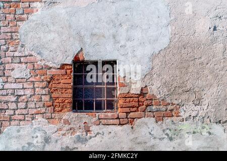 Kleines Fenster mit einem Gitter in einer alten Ziegelwand und rissigem schmutzigem Putz. Stockfoto