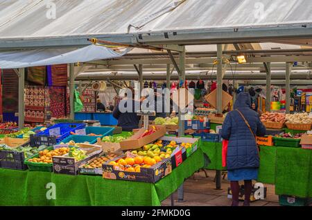 Marktstand, der Obst und Gemüse verkauft. Ein Kunde wartet darauf, bedient zu werden, und andere Stände befinden sich im Hintergrund. Stockfoto