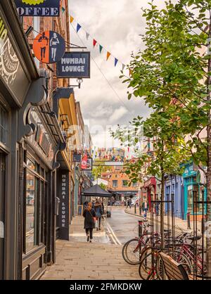 Eine Straße, die mit bunten Hahnenkästen befahren ist. Geschäfte und Restaurants befinden sich auf beiden Seiten. Fahrräder stehen im Vordergrund und eine Frau läuft auf dem Bürgersteig Stockfoto