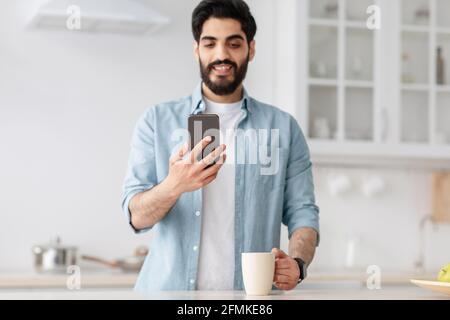 Freizeit. Junger araber liest sms auf dem Smartphone und trinkt Kaffee in der Küche. Mann genießt einen Morgentrunk, schaut sich Online-Videos an oder Stockfoto