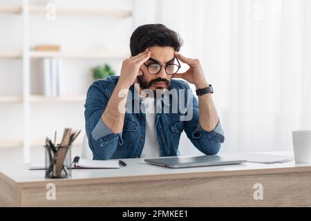 Überarbeitungs- und Terminkonzept. Gestresster arabischer Geschäftsmann, der bei der Arbeit Probleme hatte, mit einem Laptop am Arbeitsplatz saß und den Kopf berührte. Krise und Entrep Stockfoto