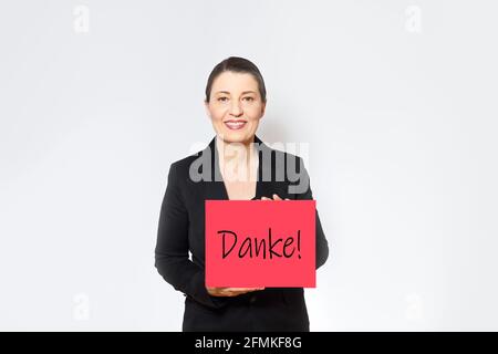 Lächelnde Frau mittleren Alters in schwarzem Blazer hält ein rotes Schild mit dem deutschen Text danke, copy space. Stockfoto