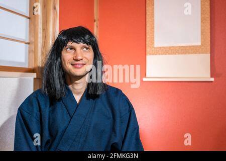 Traditionelle Schiebepapier-Shoji-Türen in Japan mit Mann in kimono Kostüm und schwarze Haare sitzen an Nische hängen Papier Scrolle lächelnd und schaue nach oben Stockfoto