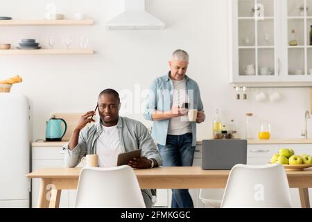 Morgens ist es voll. Reifer afroamerikanischer Kerl, der am Tablet arbeitet und am Tisch telefoniert. Leitender kaukasischer Mann tippt auf dem Smartphone und hält Tasse o Stockfoto
