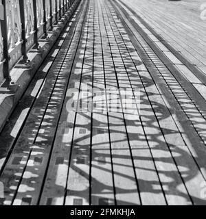 Monochrome Foto alten viktorianischen Pier und Promenade. Das Sonnenlicht strömt durch die komplizierten Designs, um Schattenmuster auf den Holzbrettern zu erzeugen Stockfoto