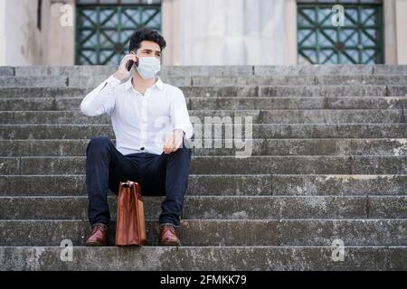 Porträt eines Geschäftsmanns, der eine Gesichtsmaske trägt und sein Mobiltelefon benutzt, während er draußen auf der Straße auf einer Treppe sitzt. Stockfoto