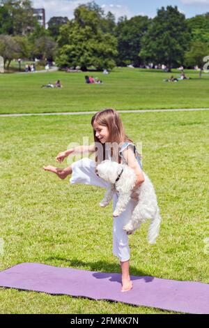 Mädchen mit Hund tut Yoga-Übungen in der Natur im Freien auf Grünes Gras auf der Fitnessmatte, Primerose Hill Park, London, Großbritannien Stockfoto