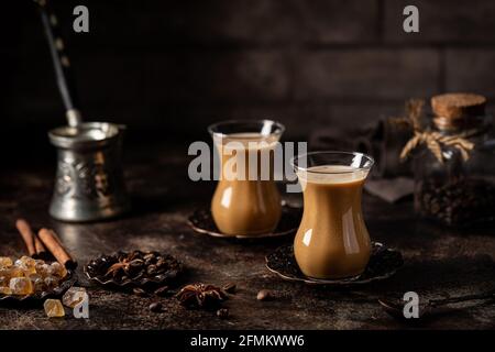 Tee oder Kaffee mit Milch und Gewürzen im Glas Armudu mit Kaffeebohnen und Zucker auf dunklem Hintergrund. Stockfoto
