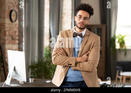Porträt eines selbstbewussten afroamerikanischen Managers mit Bart im Stehen Gekreuzte Arme gegen den Tisch mit modernem Computer im gemütlichen Büro Stockfoto