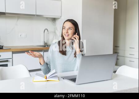 Lächelnde und freundliche junge Frau spricht auf dem Smartphone mit Laptop-Computer zu Hause, fröhliche Frau hält Telefonanruf spricht mit positivem Gesichtsausdruck, arbeitet aus der Ferne sitzen in der Küche Stockfoto