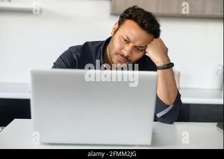 Aufgeregter multiethnischer Mann benutzt Laptop, der allein zu Hause sitzt, ein nachdenklicher junger inder sitzt in der Küche mit dem Laptop, hat mit der Hand ausgeruht, finanzielle Probleme und Probleme mit dem Projekt Stockfoto