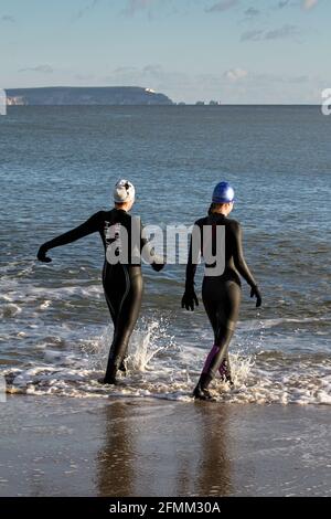 Zwei Frauen in Wetsuits, die im Winter mit Nadeln, Isle of Wight, in the Distance ins Meer gehen, um dort zu schwimmen. Christchurch Großbritannien Stockfoto