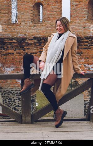 Schöne glücklich lächelnde junge Frau sitzt auf Zaun in einem beigen Mantel, weißen Wolle Kaschmir Schal und braunen Stiefeln, Street-Stil Fotografie. Filmgeräusche, so Stockfoto