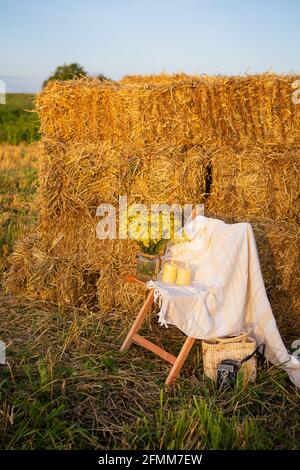 Picknick auf dem Feld in der Nähe von Strohballen. Die untergehende Sonne. Rustikaler Stil - Holzstuhl, Karo, Blumenstrauß, Kerzen Stockfoto