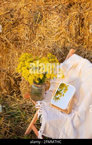 Picknick auf dem Feld in der Nähe von Strohballen. Die untergehende Sonne. Rustikaler Stil - Holzstuhl, Karo, Blumenstrauß, mirro Stockfoto