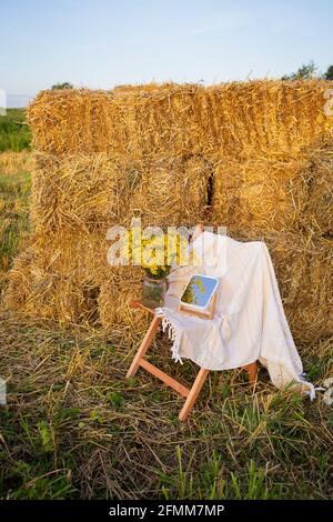 Picknick auf dem Feld in der Nähe von Strohballen. Die untergehende Sonne. Rustikaler Stil - Holzstuhl, Karo, Blumenstrauß, Spiegel Stockfoto