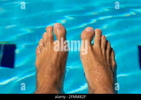 Die Füße eines jungen Mannes, der am Rand eines Pools mit kristallklarem, blauem Wasser sitzt, sich abkühlt und die Ruhe genießt, bei einer heißen Sonne Stockfoto