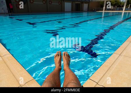 Die Füße eines jungen Mannes, der am Rand eines Pools mit kristallklarem, blauem Wasser sitzt, sich abkühlt und die Ruhe genießt, bei einer heißen Sonne Stockfoto