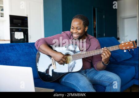 Afroamerikanischer Mann, der akustische Gitarre spielt und online streamt und aufführt, sitzt auf der Couch vor dem Laptop. Musiker gibt eine Lektion online. Schwarzer Mann lernt Gitarre spielen Stockfoto