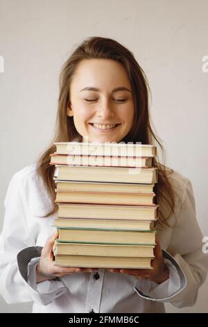 Fröhlicher junger Student, der mit geschlossenen Augen lächelt und einen großen Stapel Bücher hält. Stockfoto