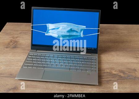 Schutzmaske auf blauem Laptop-Bildschirm, Holzschreibtisch mit schwarzem Hintergrund. Coronavirus und Antivirus-Schutz Idee. Notebook-Display mit Schnauze. Stockfoto