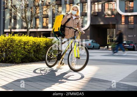 Junger Kurier in medizinischer Maske liefert Lebensmittel mit gelbem Thermalrucksack, fährt mit dem Fahrrad in der Stadt. Konzept für den Lieferservice von Lebensmitteln Stockfoto