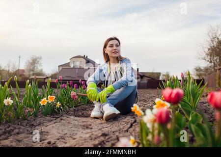 Junger Gärtner, der sich im Frühlingsgarten unter frischen Tulpen, Narzissen und Hyazinthen entspannt. Glückliche Frau genießt bunte Blumen sitzen auf dem Boden Stockfoto