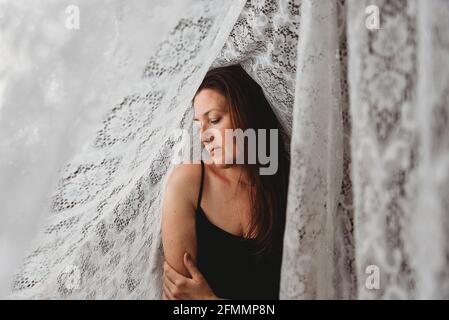 Schöne Frau mit langen braunen Haaren, umgeben von Spitzenvorhängen. Stockfoto